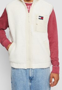 Biała kamizelka polarowa z wysokim kołnierzem, zamkiem błyskawicznym z przodu i dwoma bocznymi kieszeniami. Posiada beżową kieszeń na piersi z naszywką z logo. Nosi się ją na czerwonej koszulce z długim rękawem.