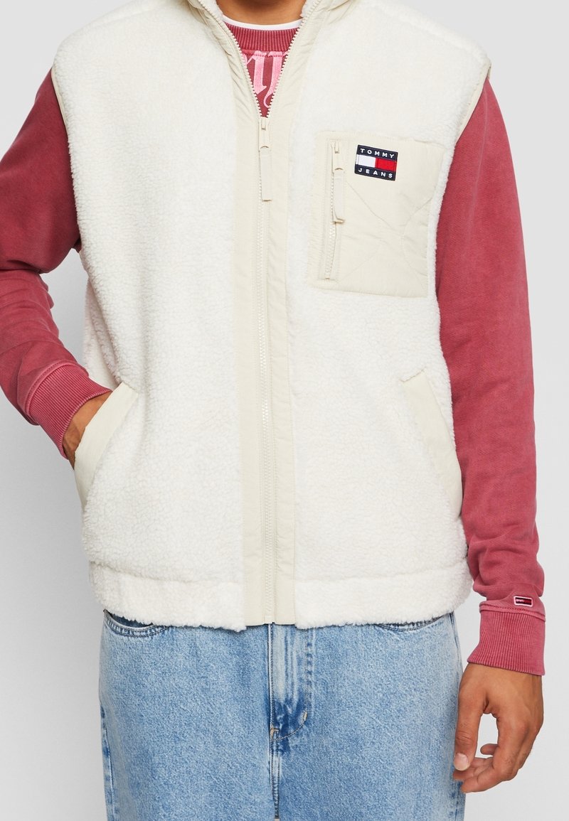 Biała kamizelka polarowa z wysokim kołnierzem, zamkiem błyskawicznym z przodu i dwoma bocznymi kieszeniami. Posiada beżową kieszeń na piersi z naszywką z logo. Nosi się ją na czerwonej koszulce z długim rękawem.