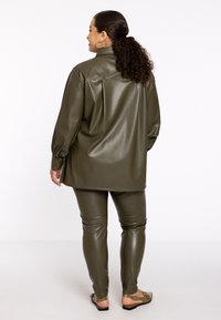 Olijfgroene faux leren shirt en broeken set met een ontspannen pasvorm. Voorzien van knoopmanchetten en een gladde textuur. G gedragen met platte sandalen.