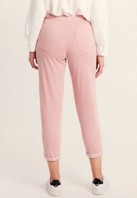 Pantalons roses fuselés avec une taille élastique et des poignets retroussés ; deux poches arrière ; tissu lisse ; associés à des baskets blanches.