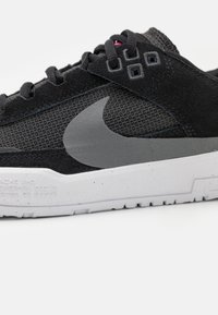 Svart joggesko laget av nett og semsket skinn med et grått Nike-logo, grå detaljer og en hvit prikkete såle. Har svarte lisser og teksturert overdel.