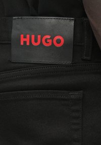 Μαύρο τζιν παντελόνι με υφασμένη δερμάτινη ετικέτα στη μέση που αναγράφει "HUGO" με έντονα κόκκινα γράμματα. Σημαντική λεπτομέρεια ραφής στις τσέπες.