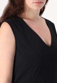 Černé šaty bez rukávů s hlubokým výstřihem do V, s hladkým povrchem a přiléhavým střihem, který zvýrazňuje čisté linie v látce.