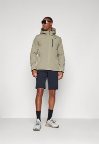 Khaki vandtæt jakke med lynlåslommer, parret med sorte shorts og lyse sneakers. Solbriller fuldender outfittet.