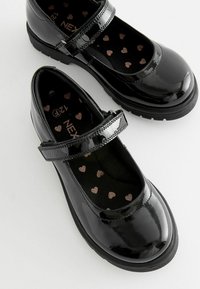 Scarpe Mary Jane in vernice nera con cinturino. L'interno presenta una fodera nera con piccoli motivi a cuore metallici. Suola piatta.