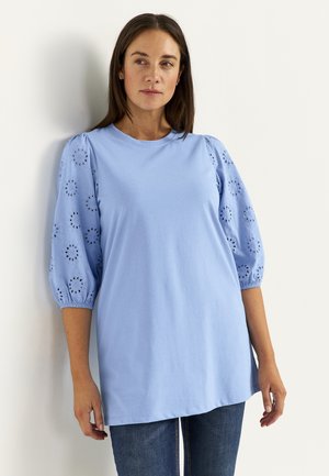 Kvinde med langt mørkt hår iført en lyseblå tunika med hulmønstrede pufærmer og blå jeans, stående op ad en hvid væg.