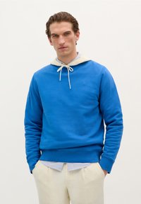 Blauwe sweatshirt met een crèmekleurige capuchon en trekkoord, gemaakt van zachte stof. Heeft ribgebreide boorden en een zoom, gedragen over een gestreept shirt.
