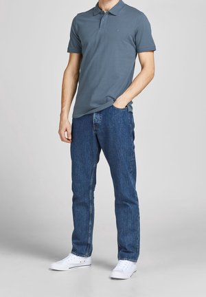 Homme portant un polo bleu, un jean bleu et des baskets blanches, debout avec une main dans la poche devant un fond uni.