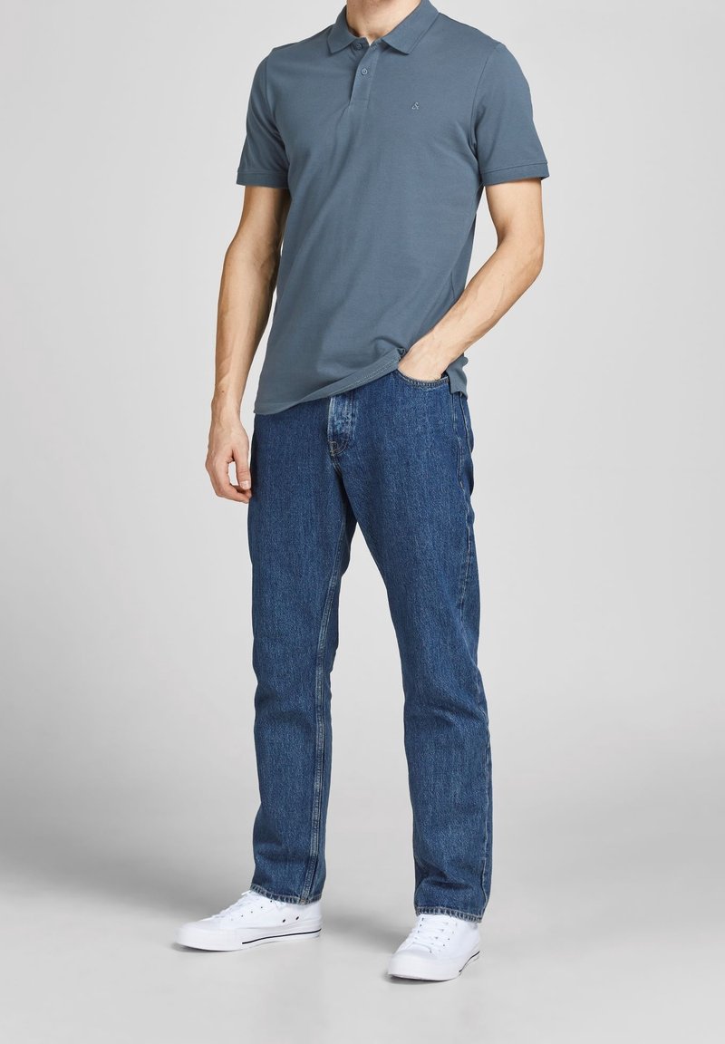 Homme portant un polo bleu, un jean bleu et des baskets blanches, debout avec une main dans la poche devant un fond uni.