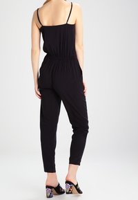 Kvinna i en svart ärmlös jumpsuit med elastisk midja, stående, kombinerad med svarta blommiga klackskor.