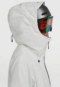 Witte waterdichte jas met een aansluitende capuchon, verstelbare trekkoorden en een rits aan de voorkant; gedragen in combinatie met een zwarte helm en kleurrijke skibril.