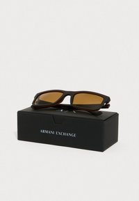 Brūnas saulesbrilles ar taisnstūra lēcām, matēta melna un brūna rāmja, atpūtoties uz melnas kastes ar uzrakstu "Armani Exchange."