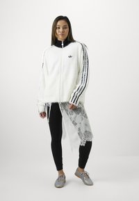 Casaco felpudo branco com colarinho preto e três riscas pretas em cada manga, combinado com leggings pretas e sapatos cinzentos. Detalhe de renda na bainha.