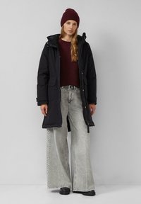 Schwarzer Parka mit Kapuze, zwei vorderen Taschen, über einem kastanienbraunen Pullover getragen, kombiniert mit weit geschnittenen hellgrauen Jeans und schwarzem Schuhwerk.