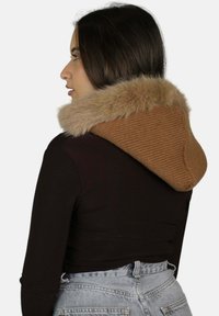 Giacca marrone a coste con cappuccio e colletto in pelliccia sintetica, abbinata a una maglia nera e jeans a vita alta azzurri chiari.