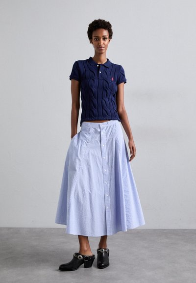Marineblaue Poloshirt aus Kabelstrick mit kurzen Ärmeln, kombiniert mit einem knielangen Rock in Hellblau und Weiß mit Streifen, ausgestattet mit Knöpfen und Taschen. Schwarze hohe Clogs.