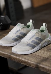 Двойка бели и сиви атлетични обувки Adidas, поставени върху дървена повърхност с черна гирица на заден план.