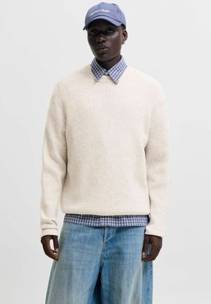 Personne portant une casquette bleue, un pull en maille blanc par-dessus une chemise à carreaux bleue, et un jean bleu clair à jambes larges, debout devant un fond uni.