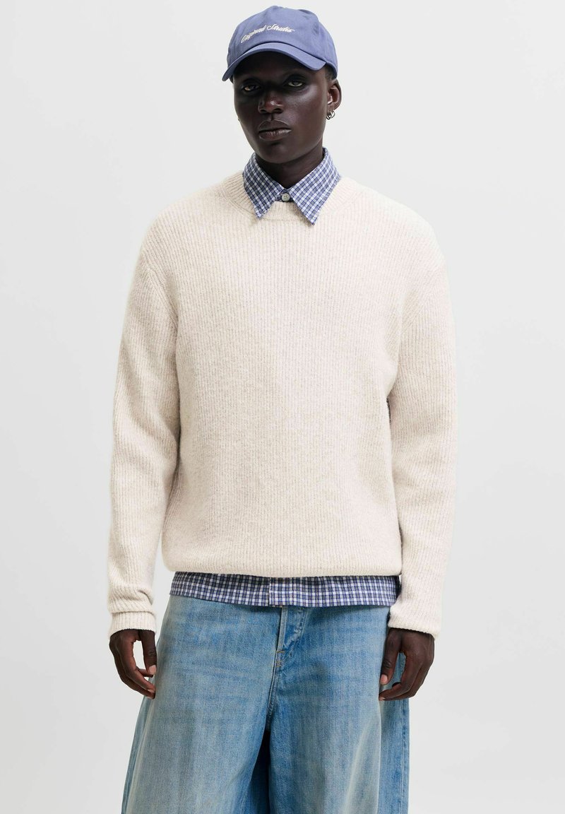 Personne portant une casquette bleue, un pull en maille blanc par-dessus une chemise à carreaux bleue, et un jean bleu clair à jambes larges, debout devant un fond uni.