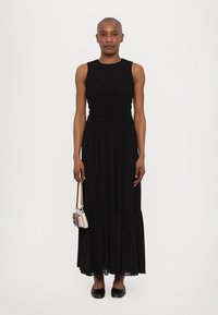 Rochie maxi fără mâneci, de culoare neagră, cu bust ajustat, fustă stratificată și material ușor. Modelul ține o mică geantă de mână cu dungi.