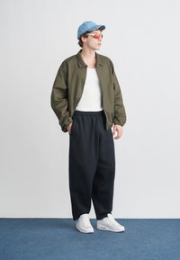 Jauns vīrietis, kurš valkā zilu cepuri, sarkani tonētas saulesbrilles, olīvu krāsas jaku, baltu topu, melnas vaļīgas bikses un baltas sporta kurpes, stāv uz zilas paklāja.