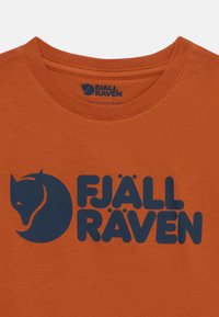 Oranžinė medvilninė marškinė su mėlynu "FJÄLL RÄVEN" logotipu ir lapės siluetu. Standartinė apvali iškirptė ir glotni tekstūra.