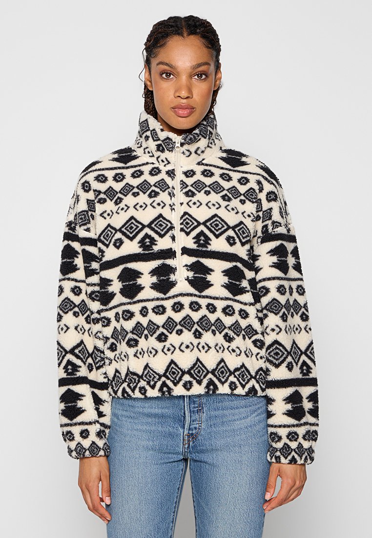 Fleece-Pullover mit hohem Kragen, cremefarbenen und schwarzen geometrischen Mustern, Reißverschluss und elastischen Bündchen, kombiniert mit hellblauen Jeans.