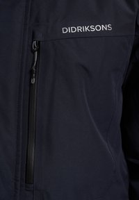 Tamsiai mėlynas Didriksons švarkas, pasižymintis užtrauktuku uždaroma kišenę, sklandžiu audinio paviršiumi ir švariomis linijomis, su subtiliu logotipu kairėje pusėje.