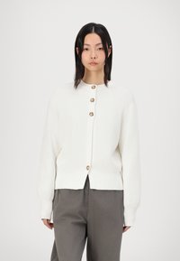 Cardigan blanc en tissu tricoté texturé, avec un col rond et cinq boutons marron sur le devant. Porté avec un pantalon gris.