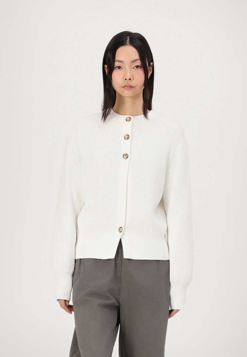 Cardigan blanc en tissu tricoté texturé, avec un col rond et cinq boutons marron sur le devant. Porté avec un pantalon gris.