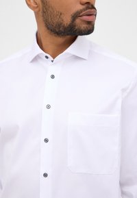 Camicia bianca con bottoni, caratterizzata da un colletto strutturato, bottoni neri e una tasca sul petto sinistro, con una finitura pulita e una texture liscia.