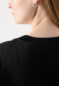 T-shirt noir à col rond et à manches courtes, avec une texture lisse. Détail visible au cou et aux boucles d'oreilles en argent avec un design géométrique.