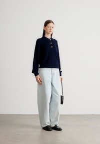 Μπλε πλεκτό πουλόβερ με ριμπ υφή, τρία κουμπιά και μυτερό γιακά, συνδυασμένο με ανοιχτόχρωμο τζιν παντελόνι φαρδιάς γραμμής και μαύρα παπούτσια.