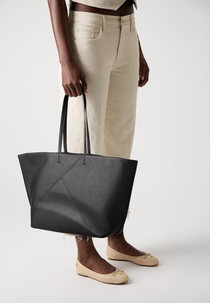 Personne portant un pantalon crème et des ballerines beiges, tenant un grand sac fourre-tout noir à longues poignées à ses côtés.