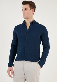 Cămașă lungă cu mâneci în culoare bleumarin, cu guler cu nasturi, dintr-un material texturat, cu croială slim fit și detalii verticale pe cusături. Asortată cu pantaloni în culori deschise.