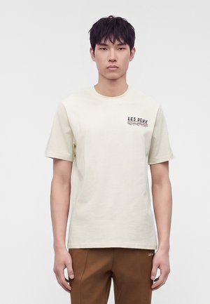 Νέος άνδρας με κοντά μαύρα μαλλιά που φοράει ένα ανοιχτό μπεζ μπλουζάκι με μικρό μαύρο κείμενο στο αριστερό στήθος και καφέ παντελόνια.