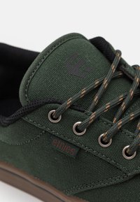 Tenis verde din pânză cu accente negre, ochiuri de metal și șireturi maro. Prezintă un logo pe limbă și o etichetă mică a brandului pe lateral.
