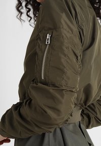 Ramię osoby noszącej oliwkową bomberkę z zamkiem błyskawicznym na rękawie i marszczonym elastycznym mankietem, w połączeniu z szarą spódnicą.