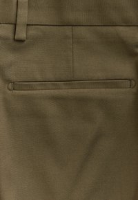 Olivinvihreä kangas, jossa on sileä herringbone-kuvio, takana yksittäinen sisätasku ja muotoiltu vyötärö.