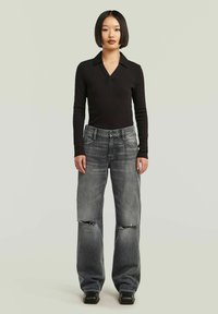Donna con capelli neri corti, che indossa un top lungo nero a maniche lunghe e jeans grigi larghi con ginocchia strappate, in piedi contro uno sfondo neutro.