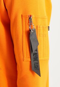 Felpa arancione con tasca a patch con zipper, accompagnata da un'etichetta in tessuto nera con marchio e testo. Dettagli testurizzati sulla tasca.