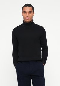 Maglione a collo alto nero a coste con maniche lunghe, caratterizzato da un design aderente e un tessuto a maglia testurizzato, abbinato a pantaloni blu navale.