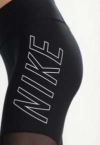 Black athletic leggings with a high waistband, featuring a prominent white "NIKE" logo vertically on the left thigh and textured mesh accents.