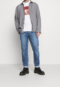 Černobílá károvaná košile, bílý grafický tričko, modré džínové kalhoty a černé kotníkové boty se žlutým prošíváním. Detaily strukturované látky.