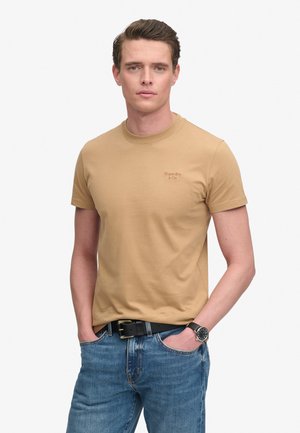 Joven con piel clara y pelo corto y marrón que lleva una camiseta beige de Superdry, vaqueros azules, cinturón negro y reloj de pulsera, manos en los bolsillos.