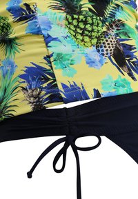 Badkleding met een gele basis, voorzien van een ananas- en bloemenpatroon in groen en blauw, gecombineerd met een marineblauwe strikbroek.