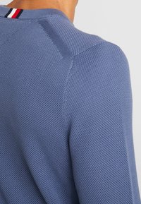 Stickad blå tröja med strukturerad yta, med en rund halsringning som accentueras av röda, vita och svarta ränder på baksidan.