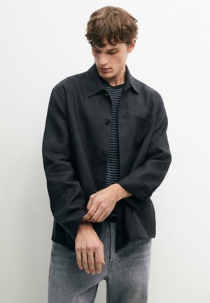 Junger Mann, der den Ärmel richtet, trägt eine schwarze Knopfleistenjacke über einem schwarz-grau gestreiften Hemd und ausgewaschene graue Jeans, steht vor einer schlichten weißen Wand.