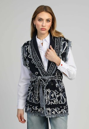 Femme portant un gilet sans manches à motifs noir et blanc avec ceinture, sur une chemise blanche boutonnée et un jean bleu clair, face à l'avant.