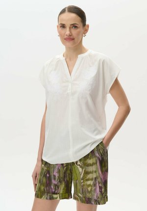 Camicia bianca a maniche corte con scollatura a V e design floreale ricamato, abbinata a pantaloni shorts verdi e viola con motivo e orlo arrotolato.